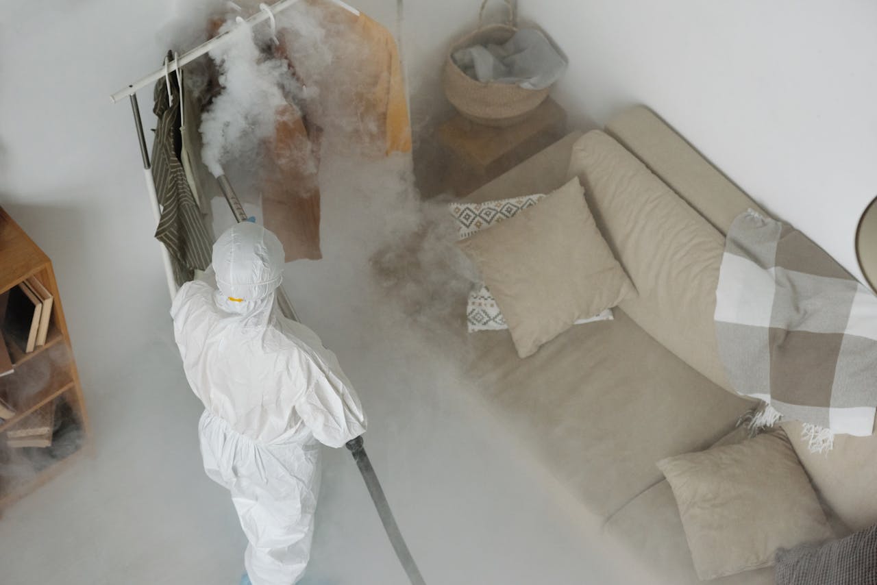 services-03 A worker in protective gear performs fumigation in a living room, using smoke to sanitize surfaces.